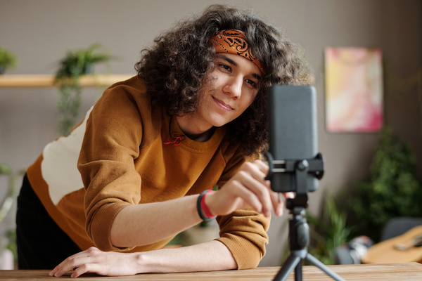 A guy with dark curly hair with a bandana in a mustard-colored sweatshirt leaning on a table is going to take a selfie on a phone that stands on a tripod in front of him